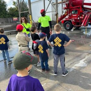 Photo of a group of youth in cub shirts and uniform. They are standing, listening to two adults, with high visibility tee shirts. They are all standing in front of a cement truck.