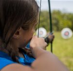 Close up, over the shoulder photo with a youth with braided hair aiming a bow and arrow to a target in the foreground