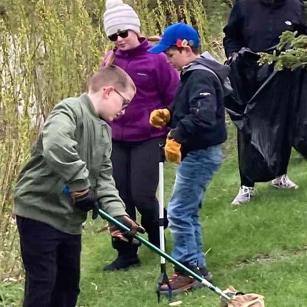 Photo of a group of youth, both male and female-presenting, wearing fall coats and holding long grabber bars. They appear to be collecting garbage from a park area.