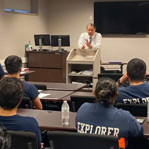 Photo of male-presenting lecturer at a podium. They are speaking to a group of youth who are sitting and listening. They are wearing dark blue shirts with the words "EXPLORER."