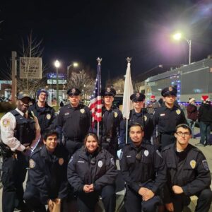 Group photo of several youth posing in two rows. It is night in downtown Joliet. The youth are all wearing police jackets and smiling. There is both an American and Illinois state flag being held by two youth in the back row.