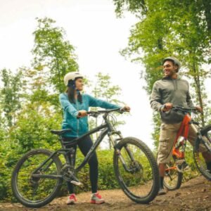An outdoor photo with two young adults posing with bicycles. They are both wearing helmets and smiling