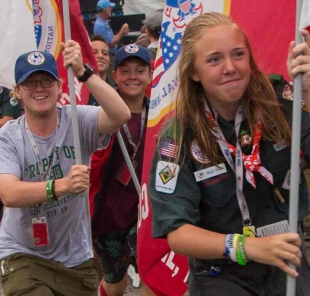 Photo of several youths holding flags. A female-presenting youth with light hair, wearing a full venturing uniform, is leading the group.