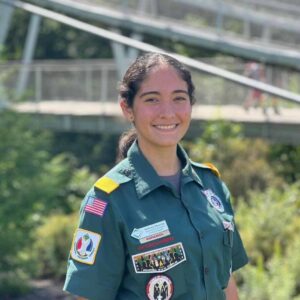 A portrait of a female-presenting person with dark hair in a green venturing uniform. They have multiple badges with one reading "Español" and they are smiling
