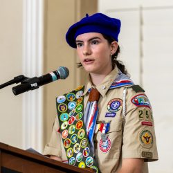 Portrait photo of female-presenting individual with long dark hair. They are wearing a tan scouting uniform with several badges and a hat. They are standing and speaking at a podium.